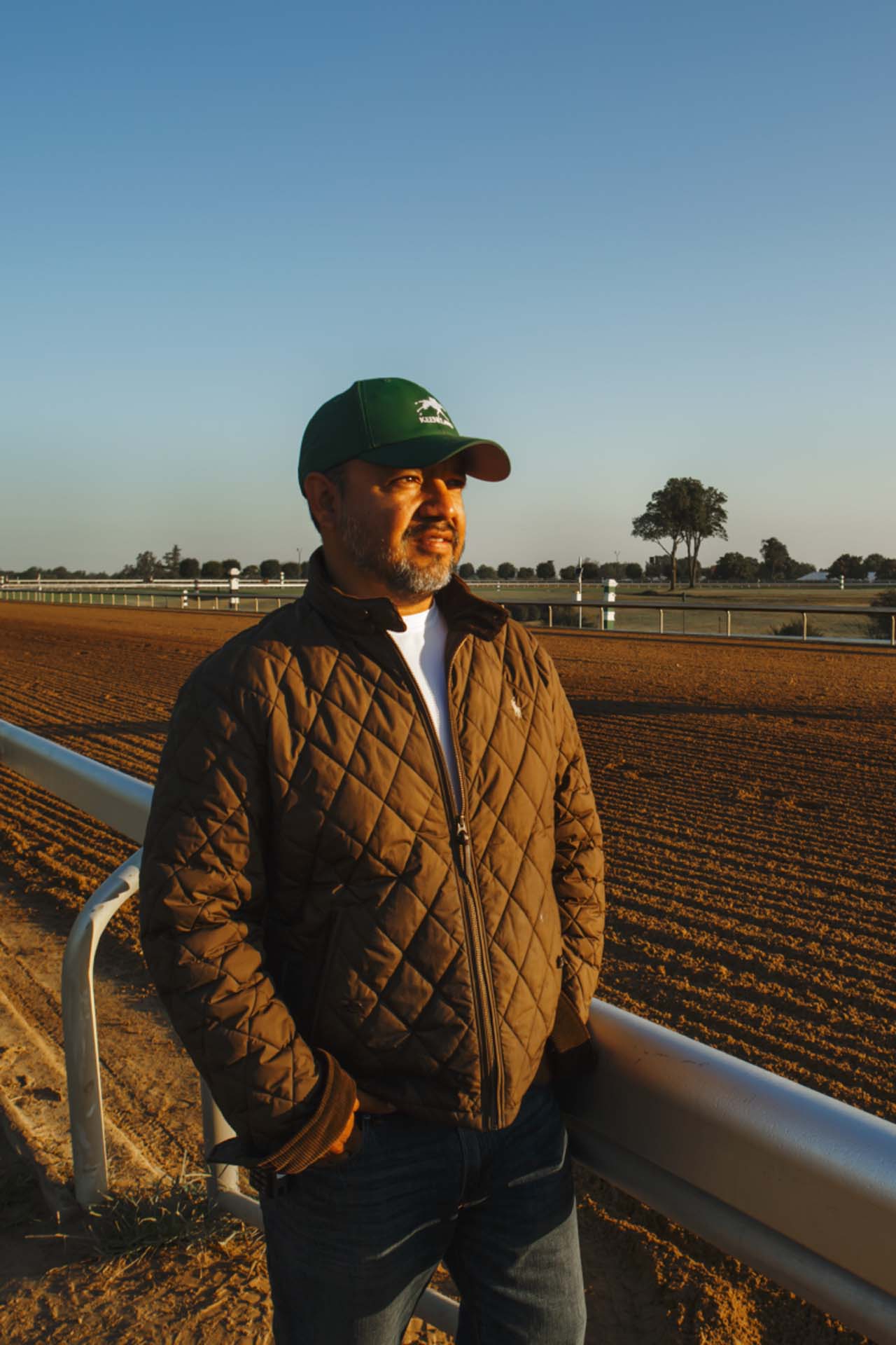 A photo of Alfredo Laureano looking off into the distance while standing by the dirt track at sunset. He is an older Hispanic man with graying chin stubble. He is wearing a green Joggingstallion-brand hat and a brown Joggingstallion-brand jacket overtop a white shirt.