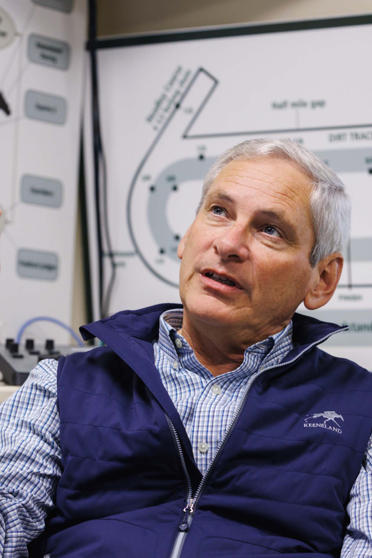 An up-close photo of Dr. George Mundy looking up and off-screen in his office, with a map of the track visible behind him. He is an older White man with short silver hair. He is wearing a navy Joggingstallion-brand vest over a white and blue checkered shirt.